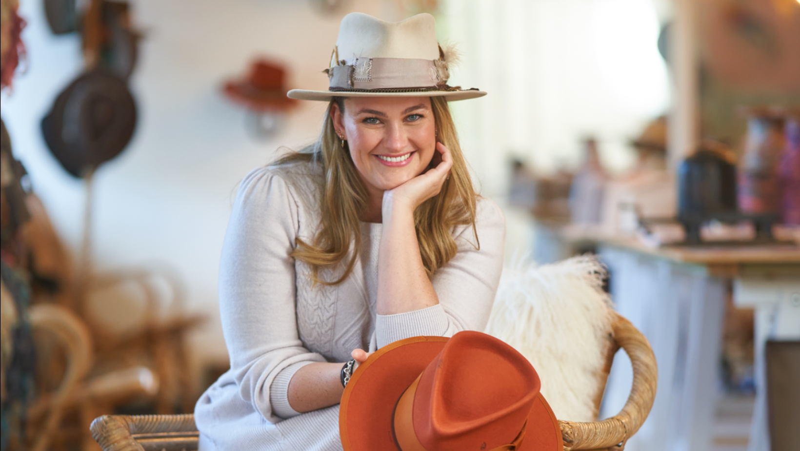 Laura Hall in her studio smiling, wearing a detailed beige hat whilst holding a hat in spiced pumpkin