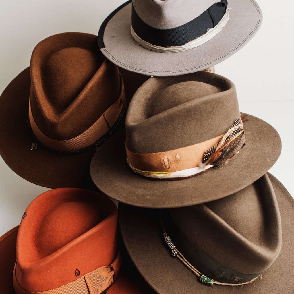 A stack of men’s handcrafted felt hats by PHYLLi Designs in earthy tones, featuring ribbon bands and unique trims