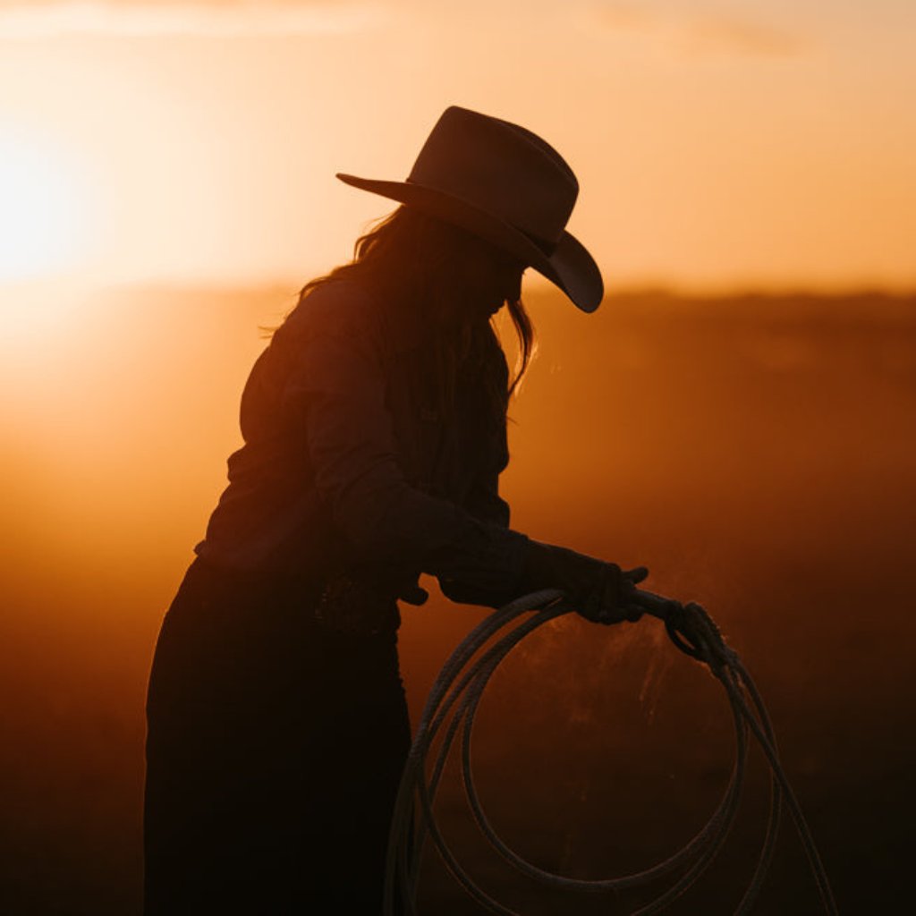 Silhouette of a woman in a Wheeler cowboy hat roping at sunset, representing the spirit of the Wheeler Collection by PHYLLi Designs