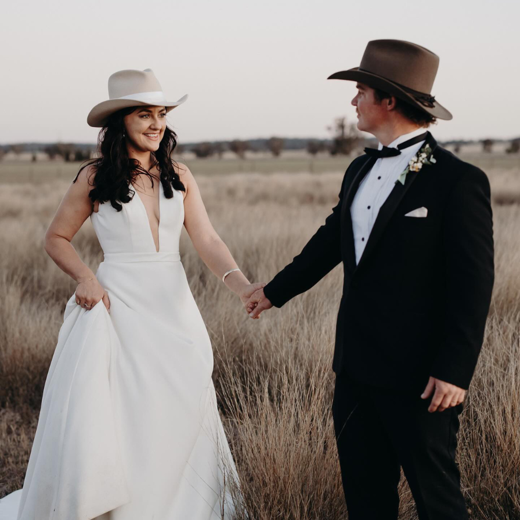 Bride and groom standing in a country field at sunset, both wearing hats from PHYLLi Designs’ Wedding Season collection - a beige felt bridal hat and a classic olive groom’s hat