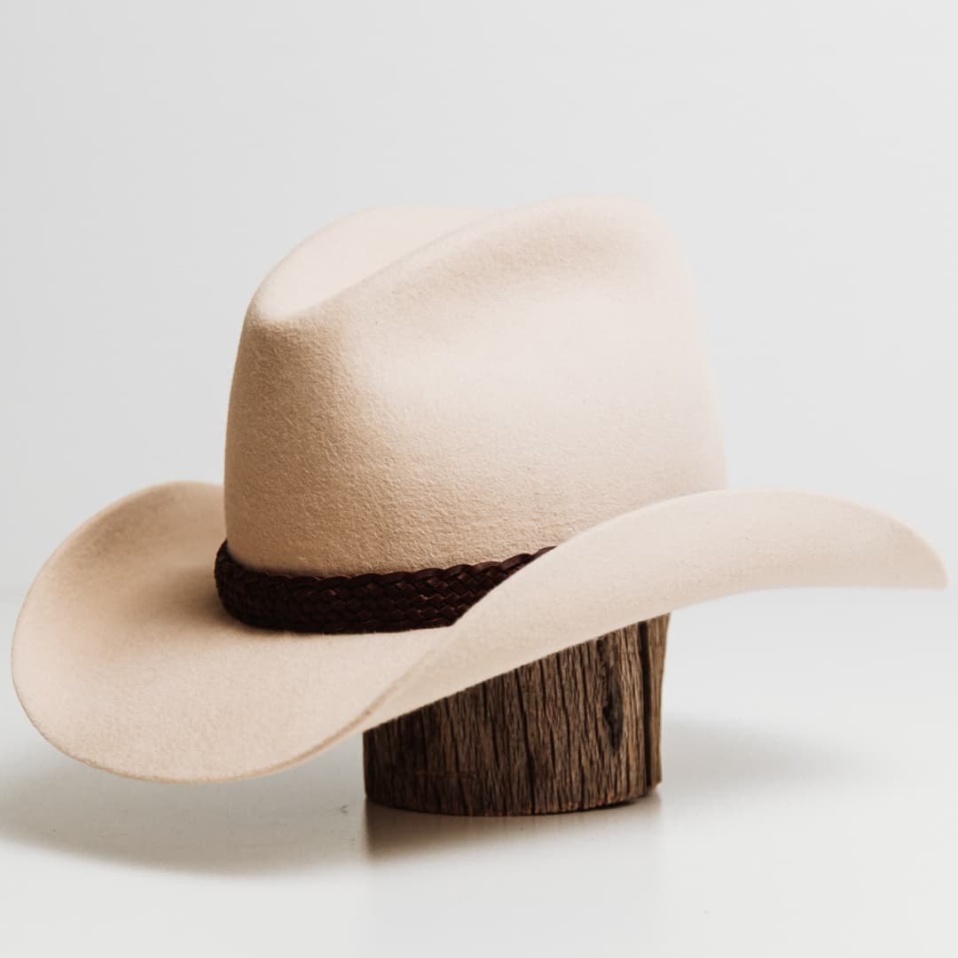 Front left angle of the 12 Strand Plaited Hat Band featuring chestnut plaited kangaroo hide with antique bronze buckle on a beige felt cowboy hat by PHYLLi Designs.