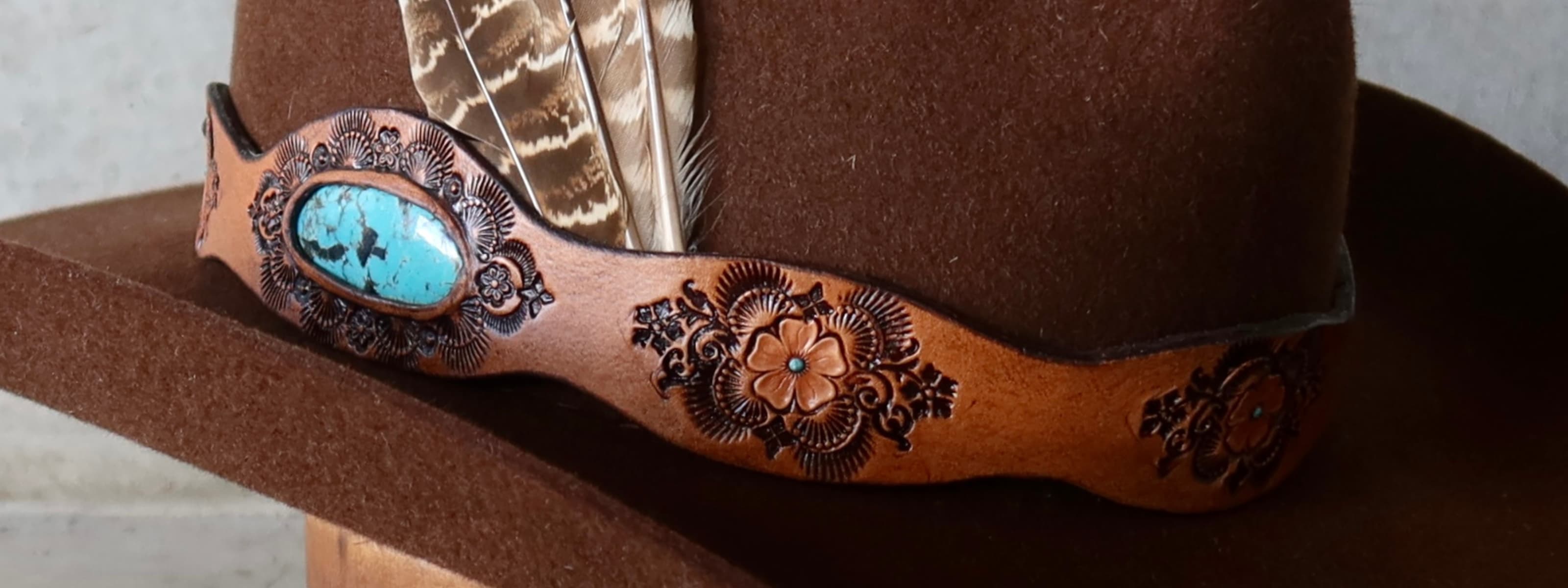 Brown cowboy hat with decorative band featuring turquoise stone and feathers.