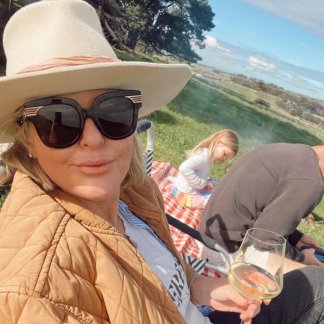 Emma Hawkins in a hat and sunglasses holding a glass of wine outdoors with people in the background.