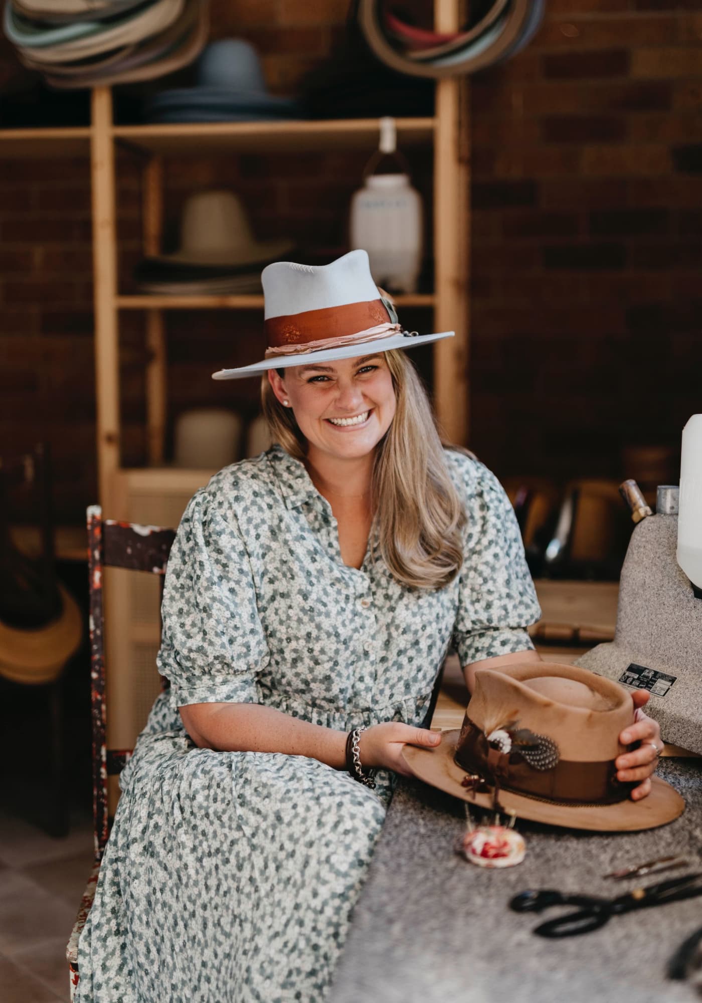 Laura Hall wearing a blue hat and holding another hat in a rustic setting with shelves of hats.