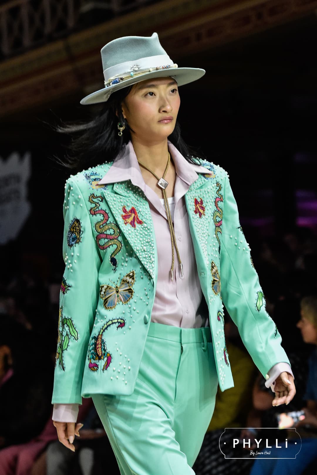 Woman wearing the Accra sage felt hat by PHYLLi Designs with cornflower bow and sari silk trimming walking on a runway in mint embroidered suit.