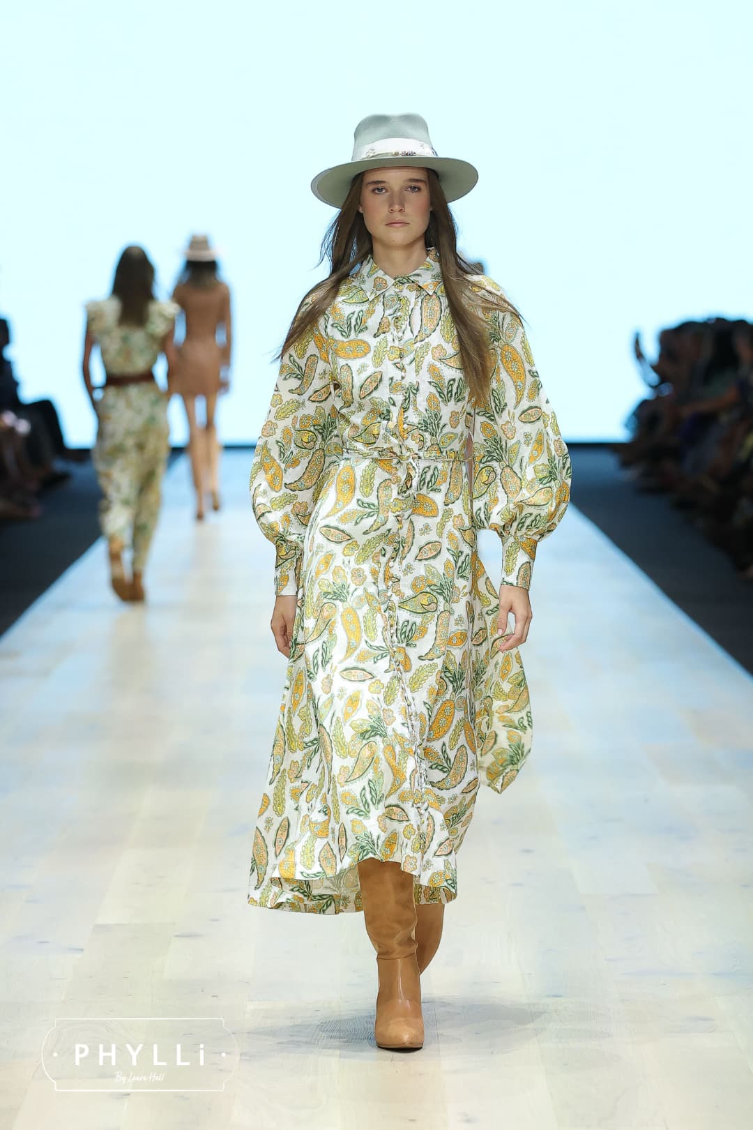 Woman wearing the Accra sage felt hat by PHYLLi Designs with sari silk trimming and Ghanaian glass bead detailing walking on a fashion runway.