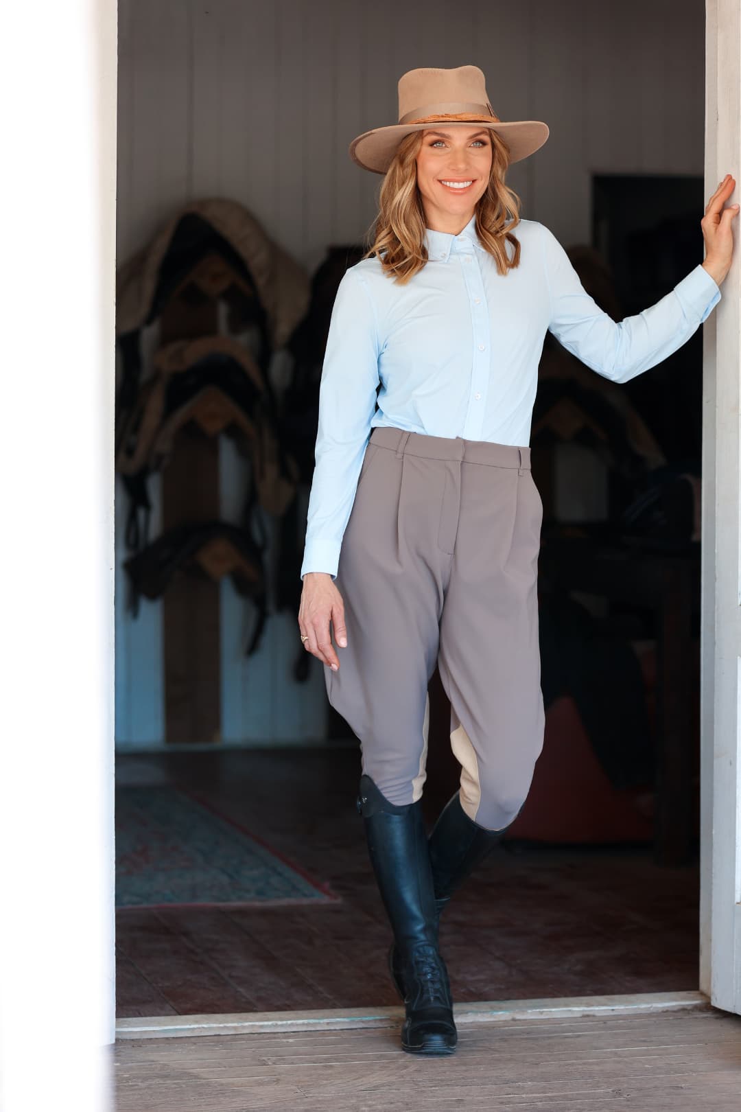 Woman wearing the Alexander Camel felt hat by PHYLLi Designs with taupe grosgrain band while standing in a stable doorway in equestrian attire.