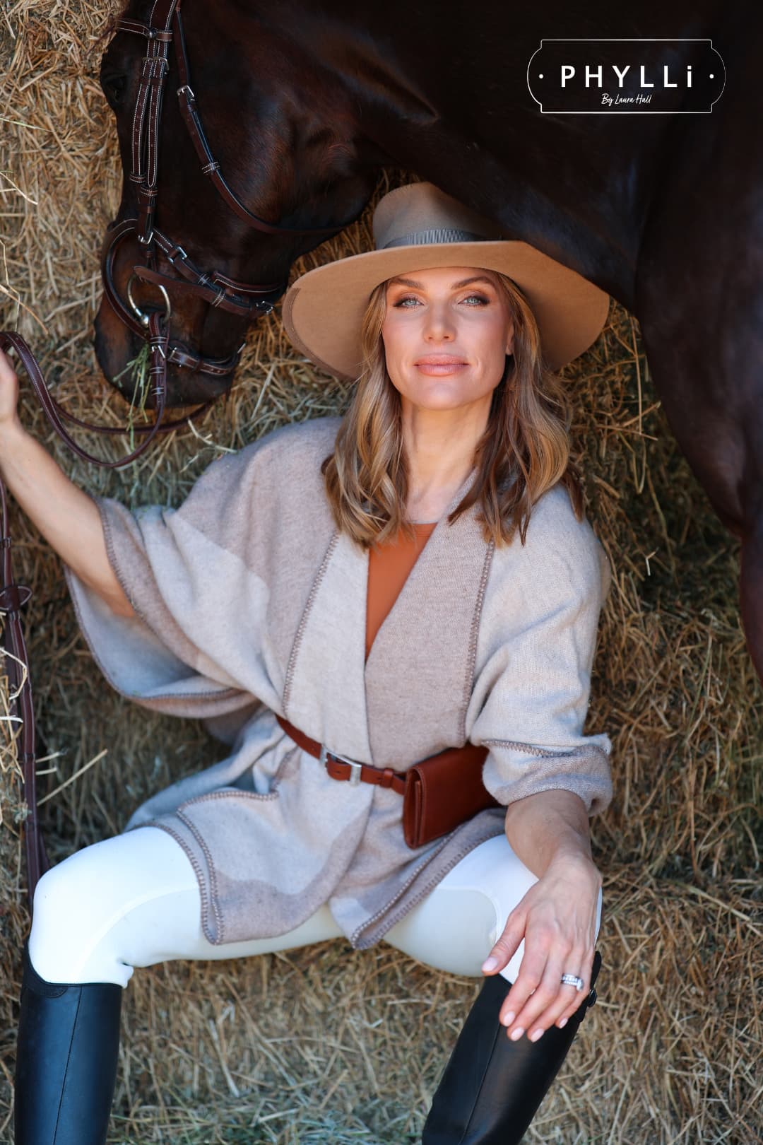 Woman wearing the Alexander Camel felt hat by PHYLLi Designs with chocolate suede string detail while sitting beside a horse in a barn.