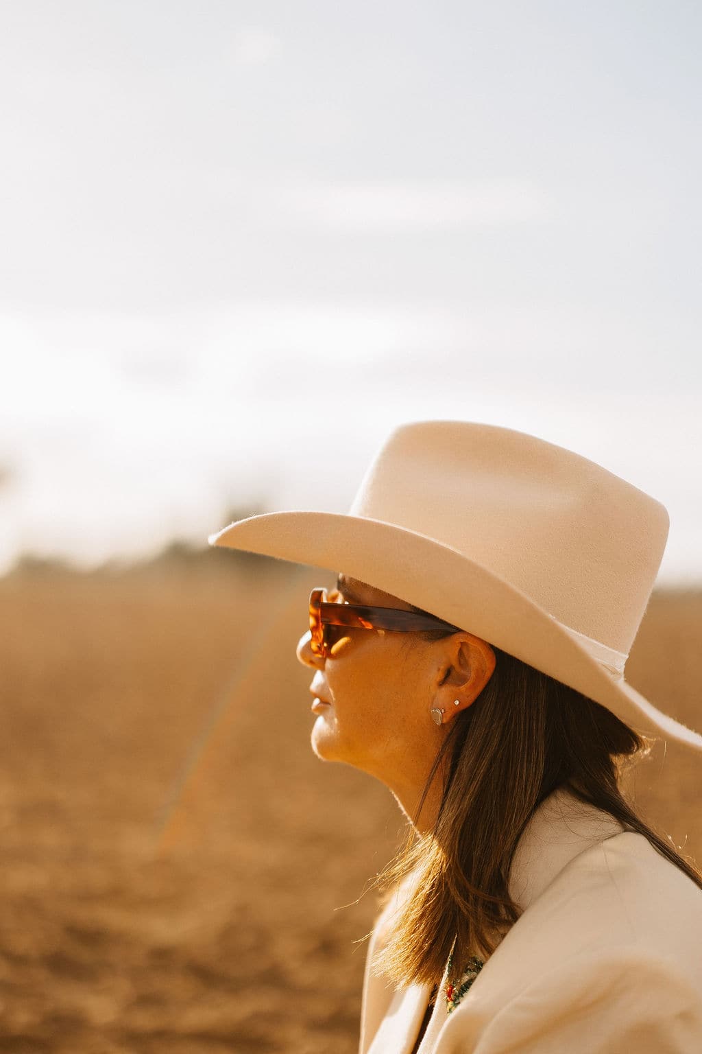 Side profile of a woman wearing the PHYLLI B&W Wheeler beige cowboy hat and sunglasses in golden afternoon light.