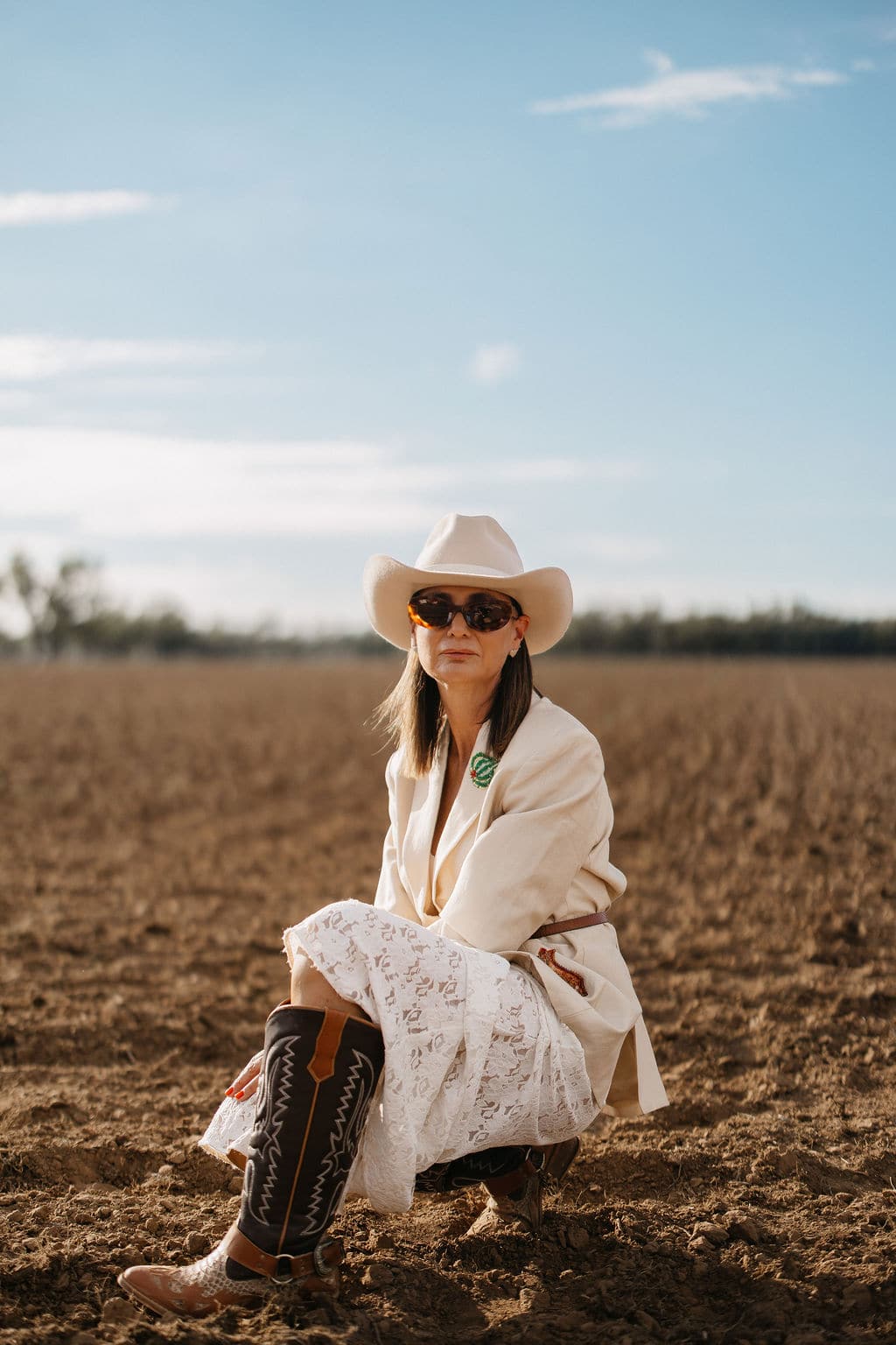 Woman wearing the PHYLLi B&W Wheeler beige cowboy hat and a linen jacket, kneeling in a paddock at sunset.
