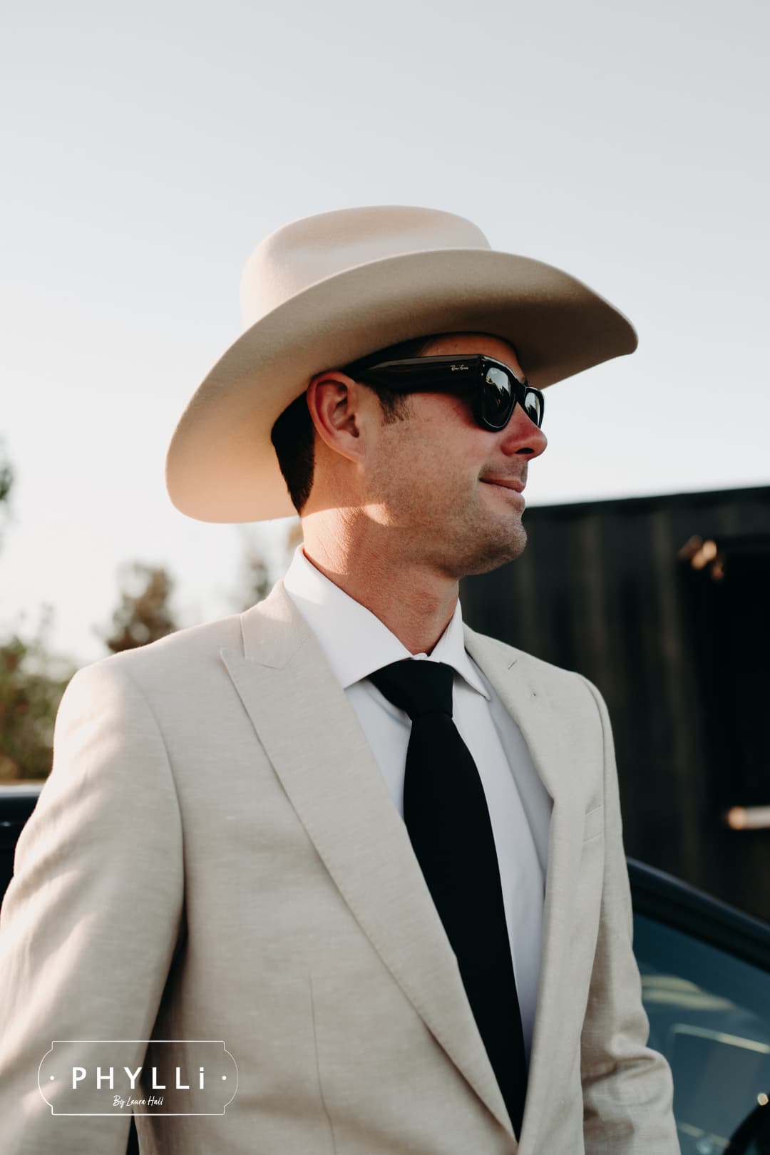 Male model wearing the beige felt cowboy hat B&W Wheeler by PHYLLi Designs with ivory Japanese grosgrain band in an outdoor event setting.