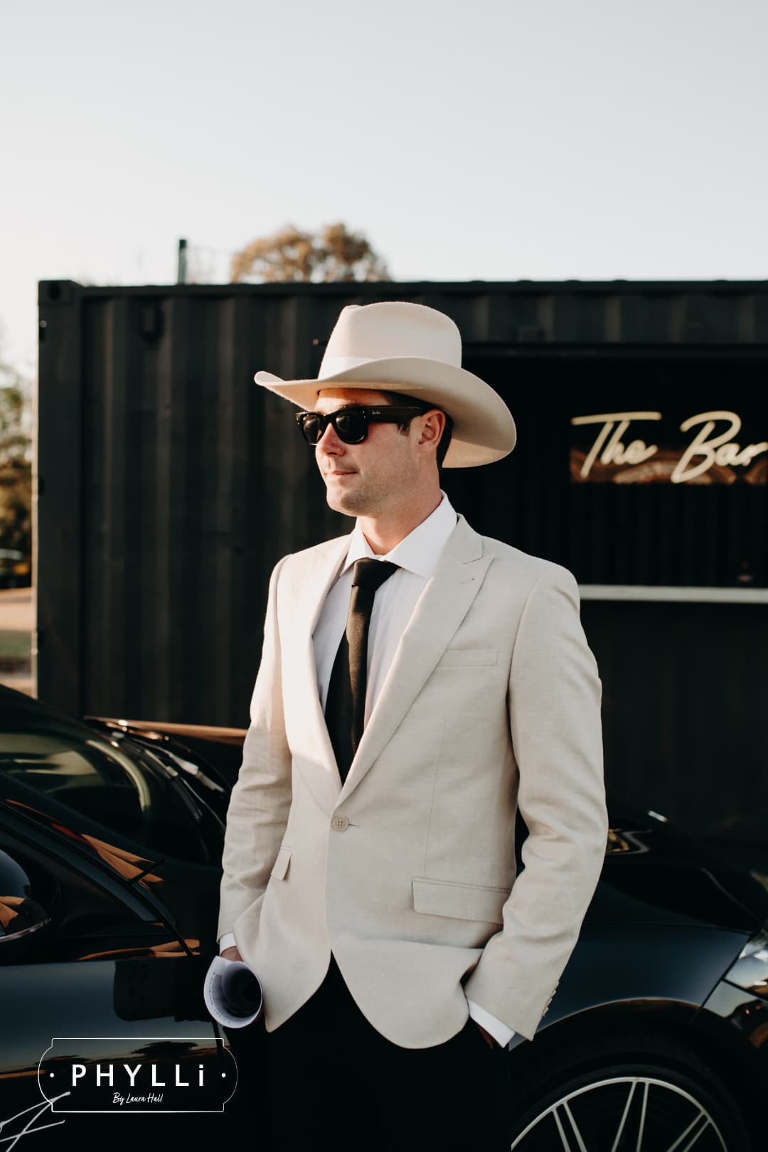Male model wearing the beige felt cowboy hat B&W Wheeler by PHYLLi Designs with ivory Japanese grosgrain band standing beside a car at sunset.
