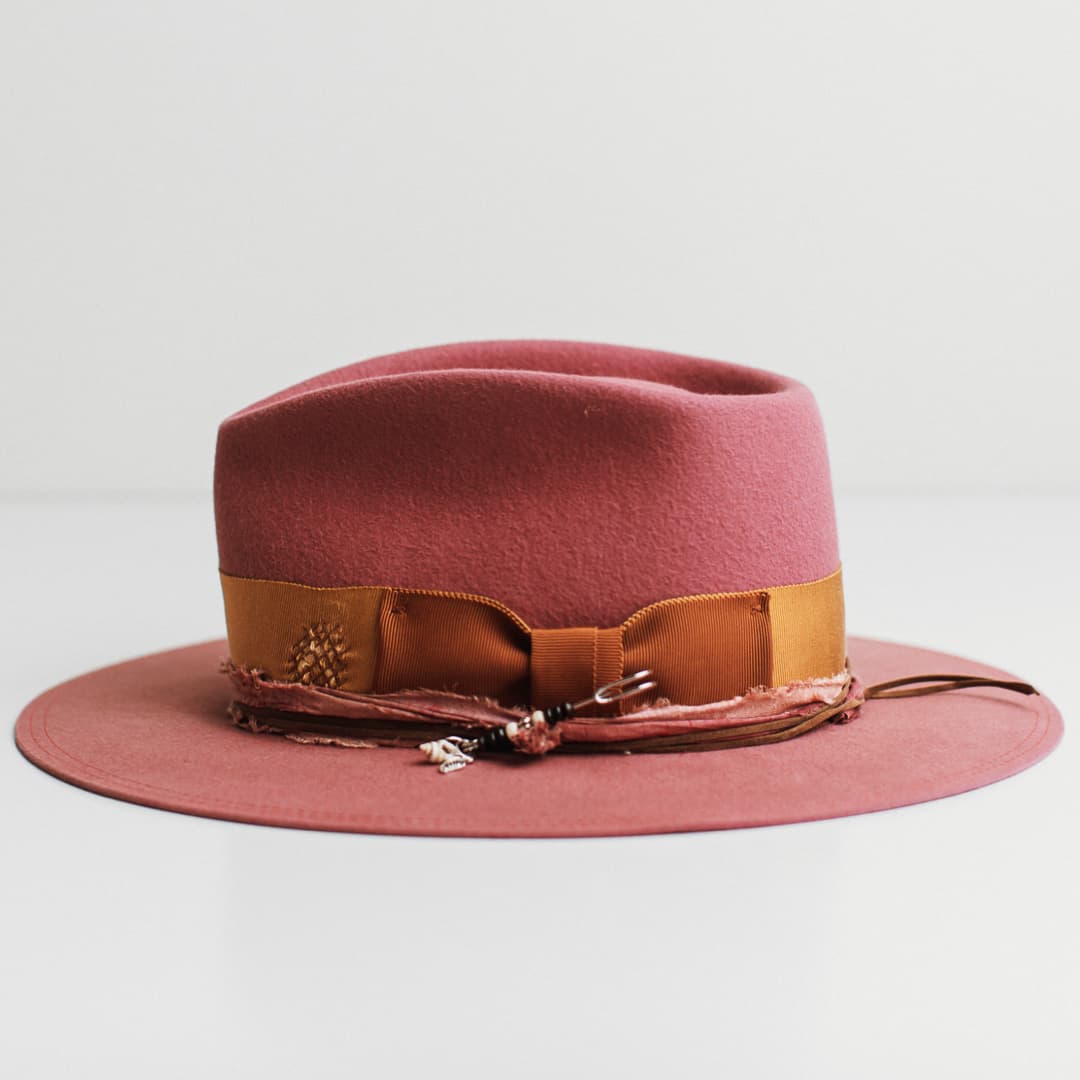 Left side view of the Barbados Rose rose felt hat by PHYLLi Designs with turmeric grosgrain band, rust bow and silver hat pin with wooden beading, shell and pendant charm.