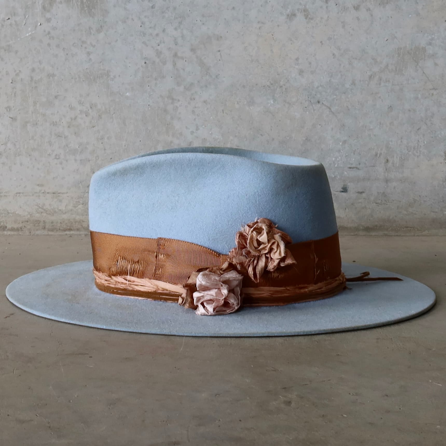 Left side view of the crystal blue felt hat Blue Bloom by PHYLLi Designs with copper grosgrain band, tan suede string wrap and layered sari silk Lapel Blooms.