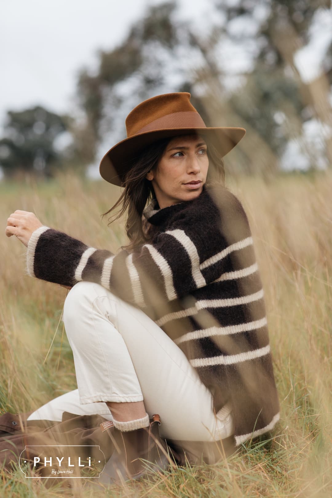 Woman wearing the Calaveras caramel felt hat by PHYLLi Designs with chocolate grosgrain band in grassy paddock setting.