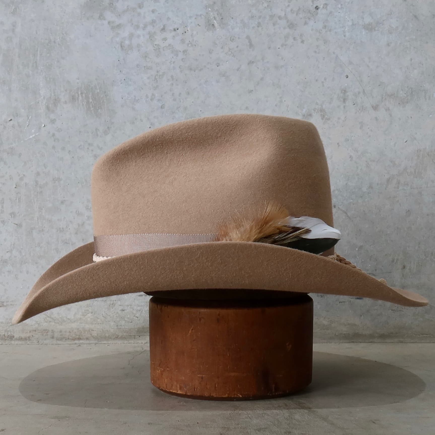 Left side view of the camel felt cowboy hat Casey by PHYLLi Designs with assorted fallen feathers and taupe grosgrain band.