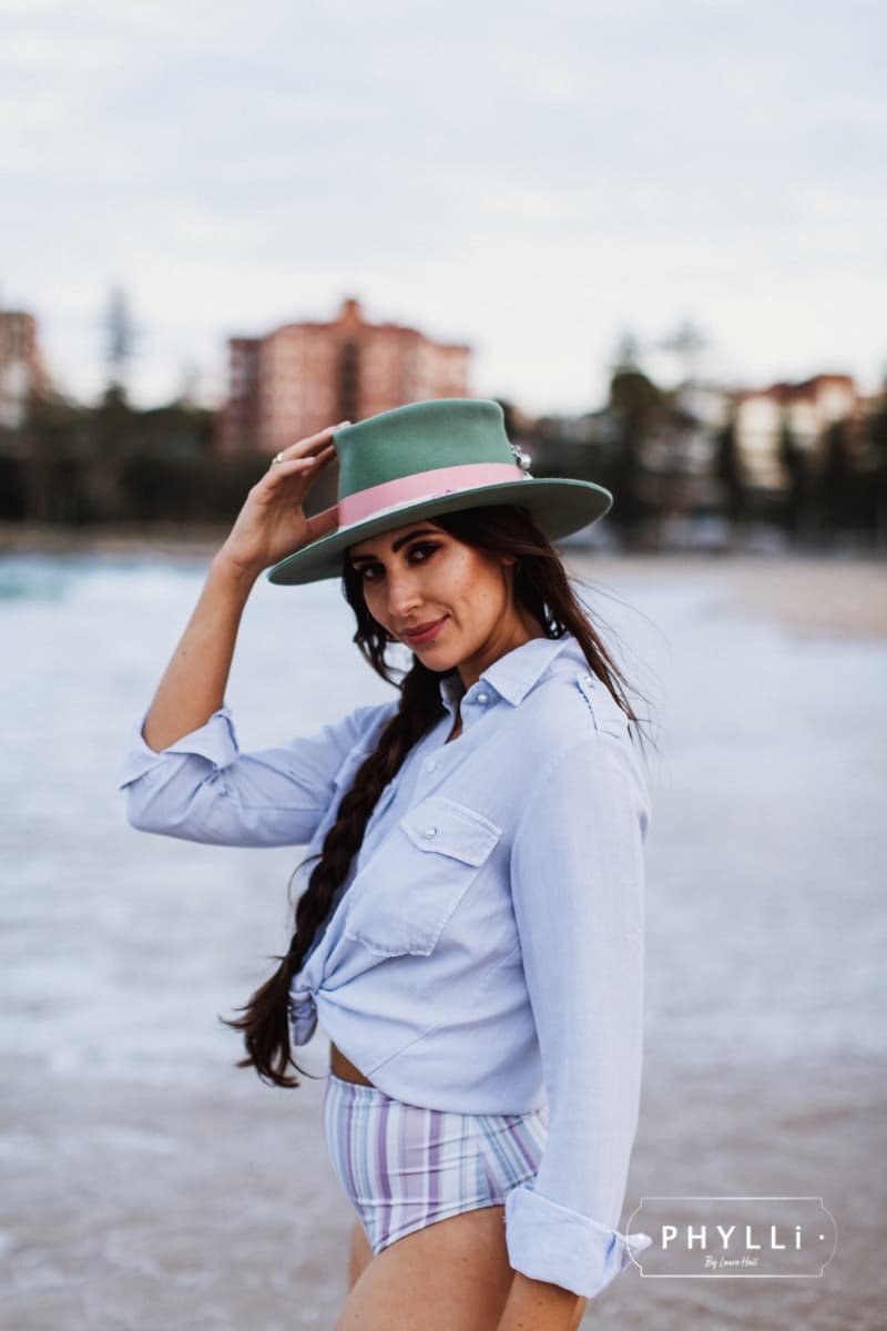 Woman wearing the Diamonds & Pearls eucalyptus felt hat by PHYLLi Designs with pearl beaded chunky silver hat pin and pink grosgrain band by the ocean.