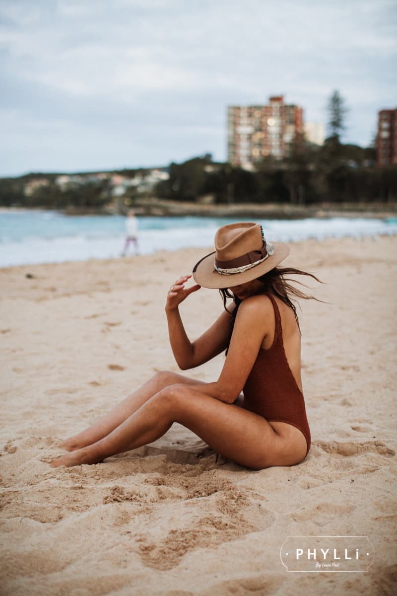 Woman wearing Fly Fisher camel felt hat by PHYLLi Designs sitting on a sandy beach in a rust swimsuit with ocean in the background.