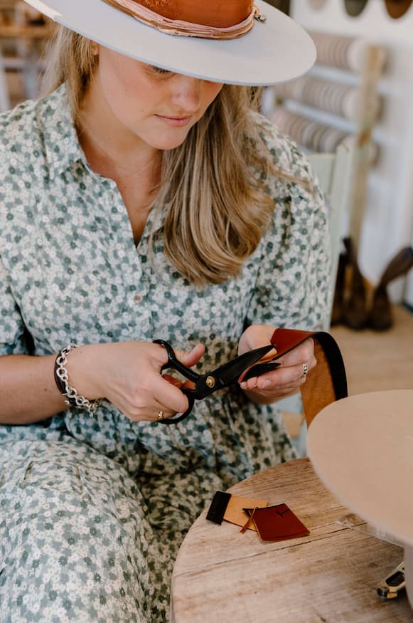 Close up detail of PHYLLi designs hatmaker Laura Hall cutting leather hatband material with scissors while wearing a felt hat in a studio setting.