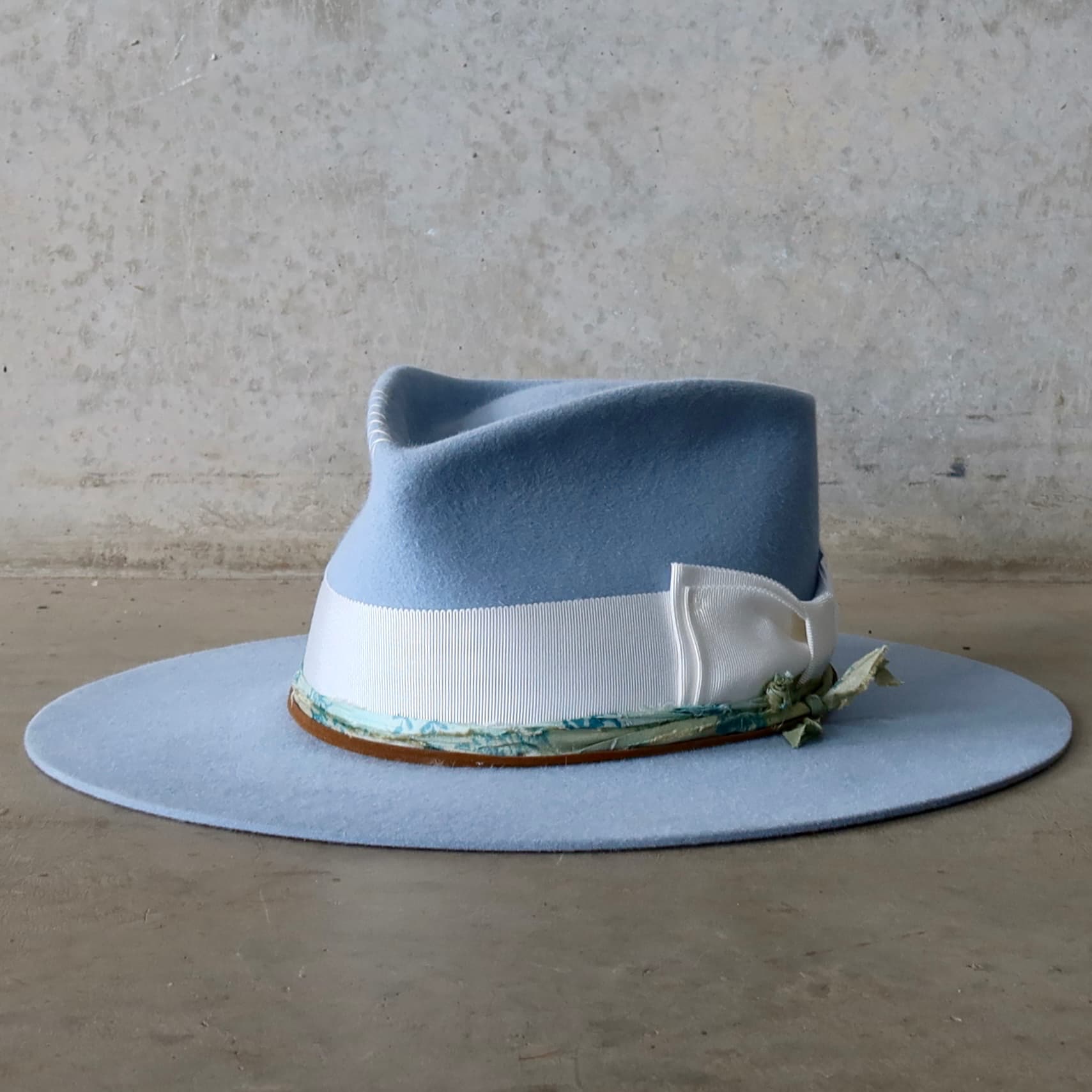 Front left angle of the crystal blue felt hat Jarvis by PHYLLi Designs with ivory grosgrain bow band, sari silk trim and tan suede string.