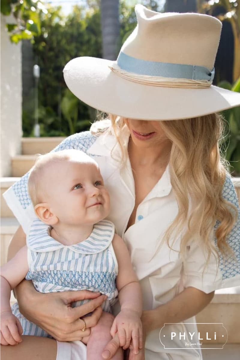 Female model wearing the beige felt hat Kingston by PHYLLi Designs with Cornflower Japanese grosgrain bow band and ivory sari silk trim holding a baby in a garden setting.