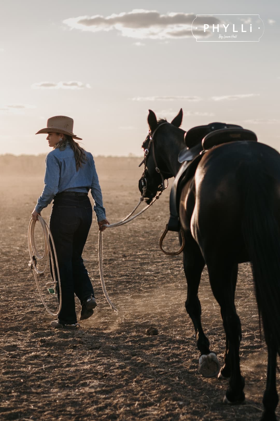 Cowgirl wearing the camel felt cowboy hat La Fleurs by PHYLLi Designs with floral burnt branding while leading a horse across a dusty paddock at sunset.