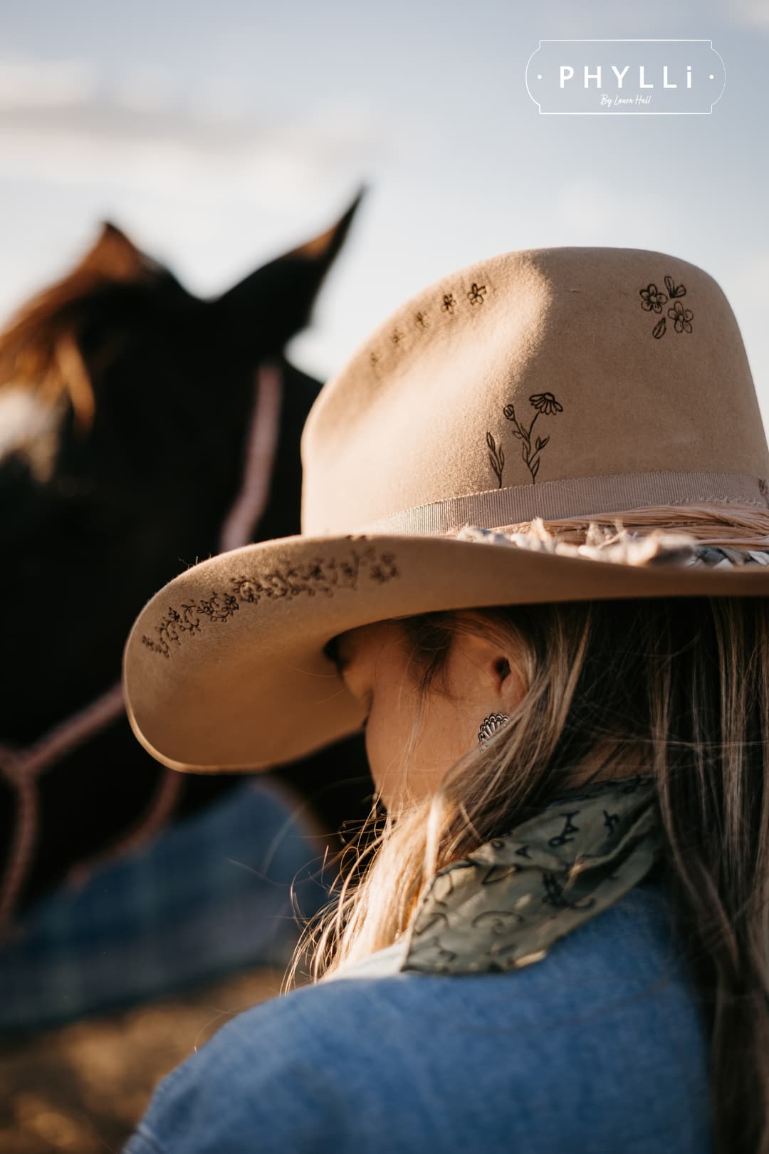 Cowgirl wearing the camel felt cowboy hat La Fleurs by PHYLLi Designs with botanical burnt branding beside a horse at golden hour.
