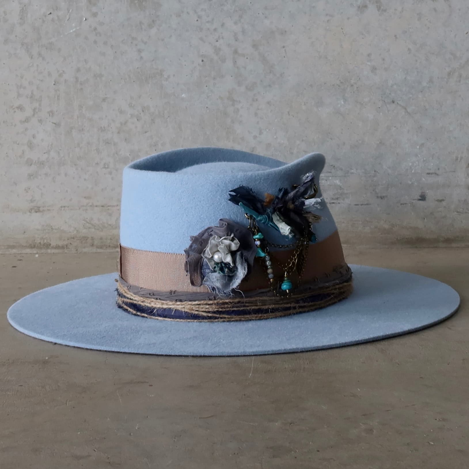 Back right angle of the crystal blue felt hat Louise by PHYLLi Designs showcasing the layered sari silk Lapel Blooms and bronze chunky hat pin with turquoise beads and draped chains.