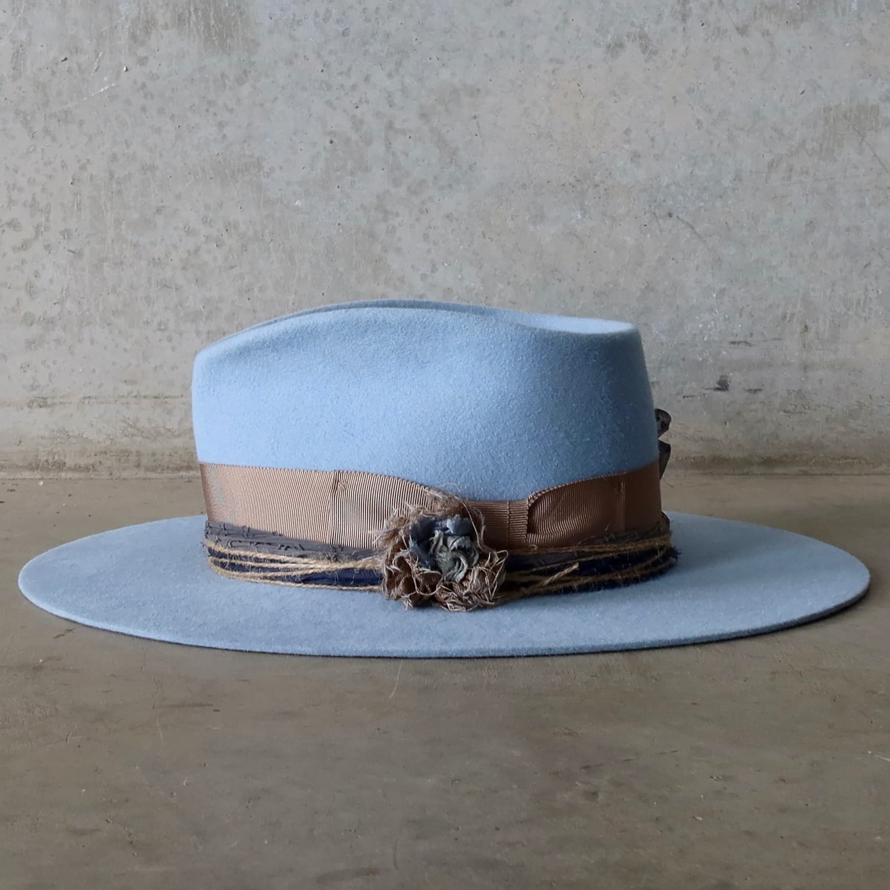 Left side view of the crystal blue felt hat Louise by PHYLLi Designs featuring taupe Japanese grosgrain band, sari silk Lapel Bloom and four twine wraps around the crown.