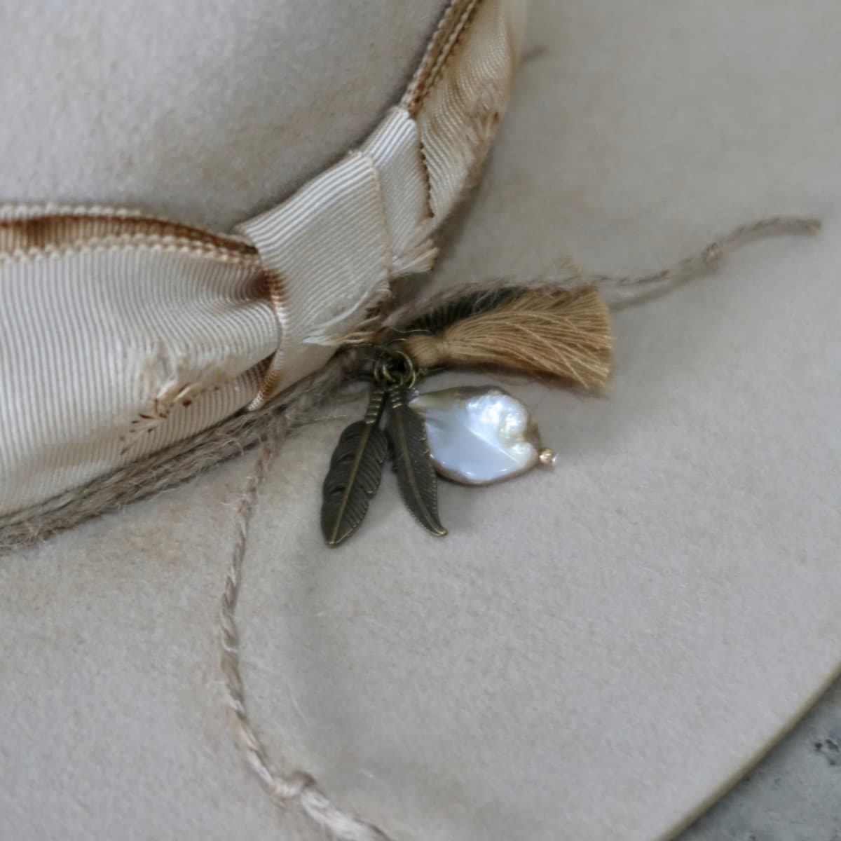 Close up detail of the Lucille Limited Edition felt hat showing bronze feather charms, tassel and rough pearl pendant.