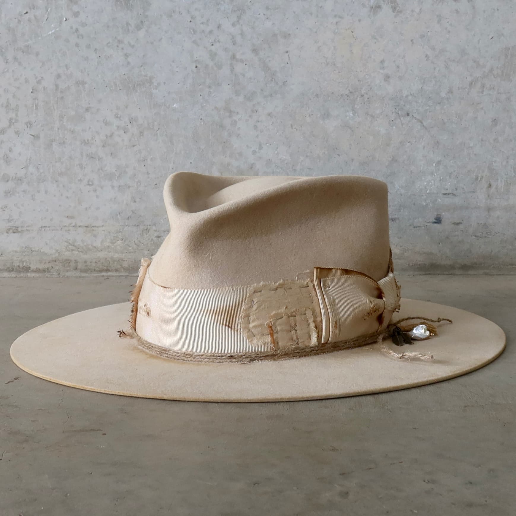 Front left angle of the beige felt hat Lucille Limited Edition by PHYLLi Designs with distressed ivory Japanese grosgrain double bow band and charm cluster featuring bronze feathers, tassel and rough pearl pendant.