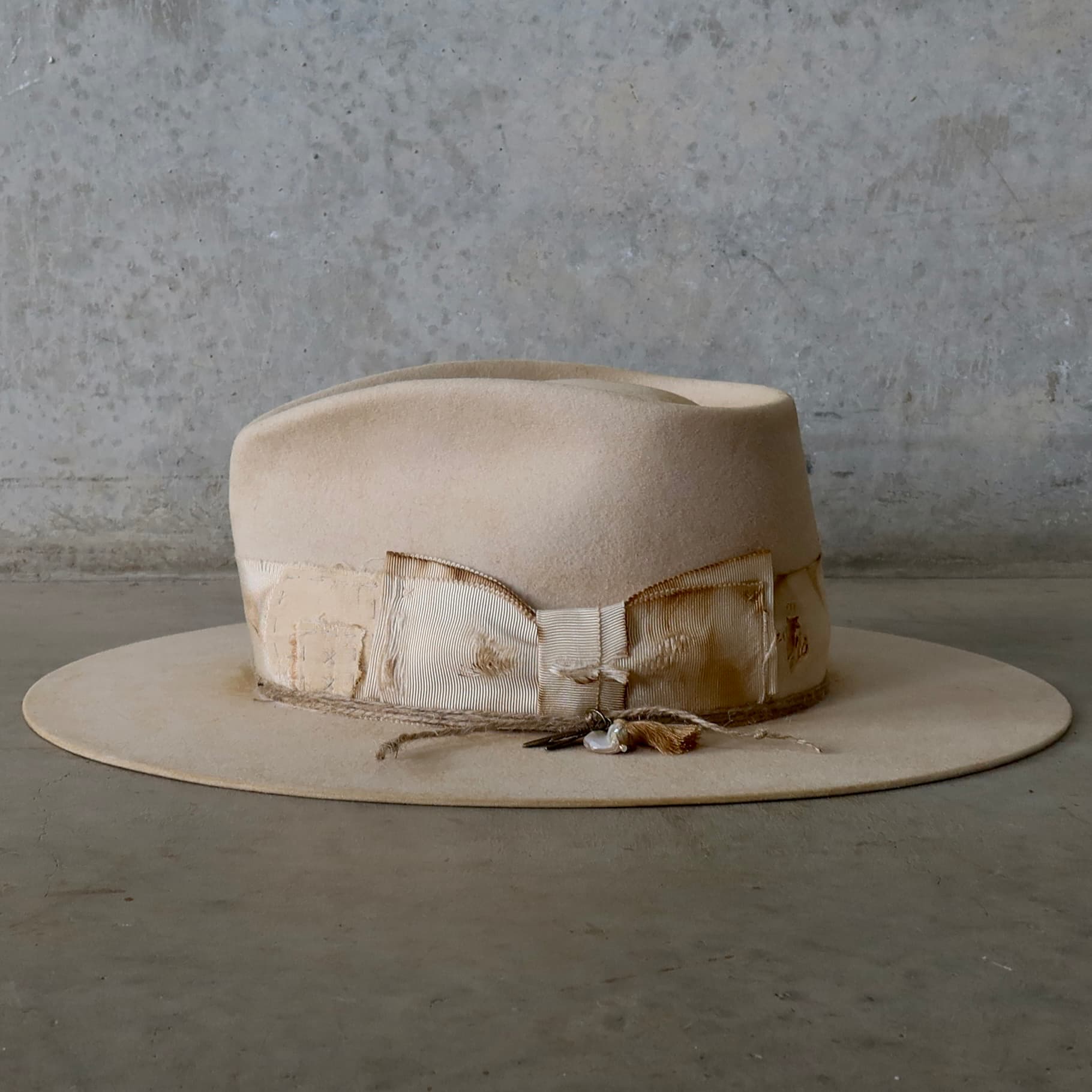 Left side view of the beige felt hat Lucille Limited Edition by PHYLLi Designs with twine wraps and charm cluster featuring tassel and rough pearl.