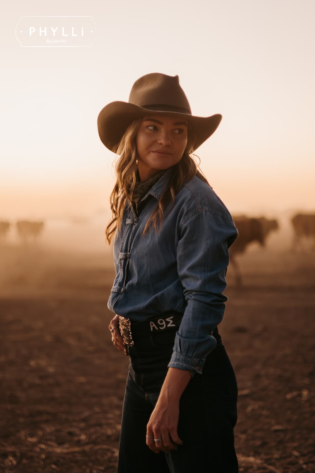 Woman wearing the Midnight Cowboy dirty olive felt cowboy hat by PHYLLi Designs with black Japanese grosgrain ribbon band and buffalo tieback hat band in dusty cattle yards at sunset.