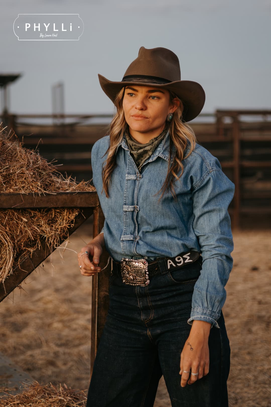 Woman wearing the Midnight Cowboy dirty olive felt cowboy hat by PHYLLi Designs with black Japanese grosgrain ribbon band and buffalo tieback hat band beside a hay bale in rural cattle yards.