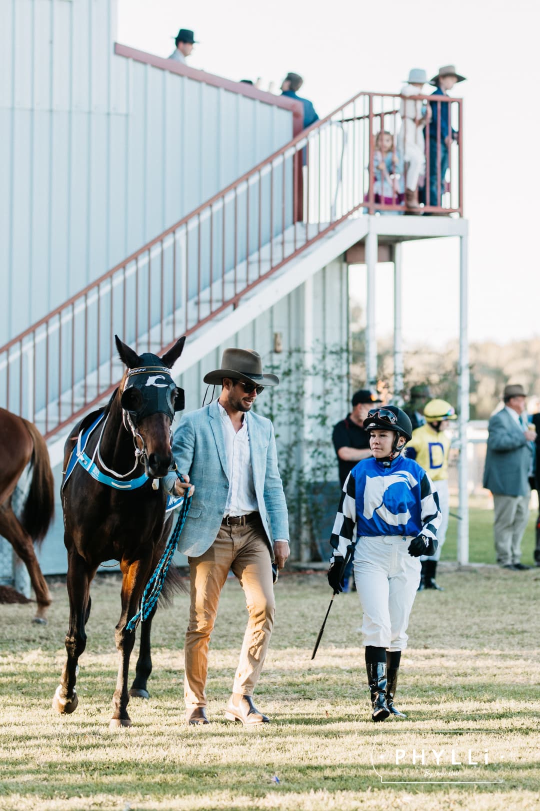 Man wearing a PHYLLi Designs felt hat while walking a racehorse beside a jockey in a racetrack parade yard.