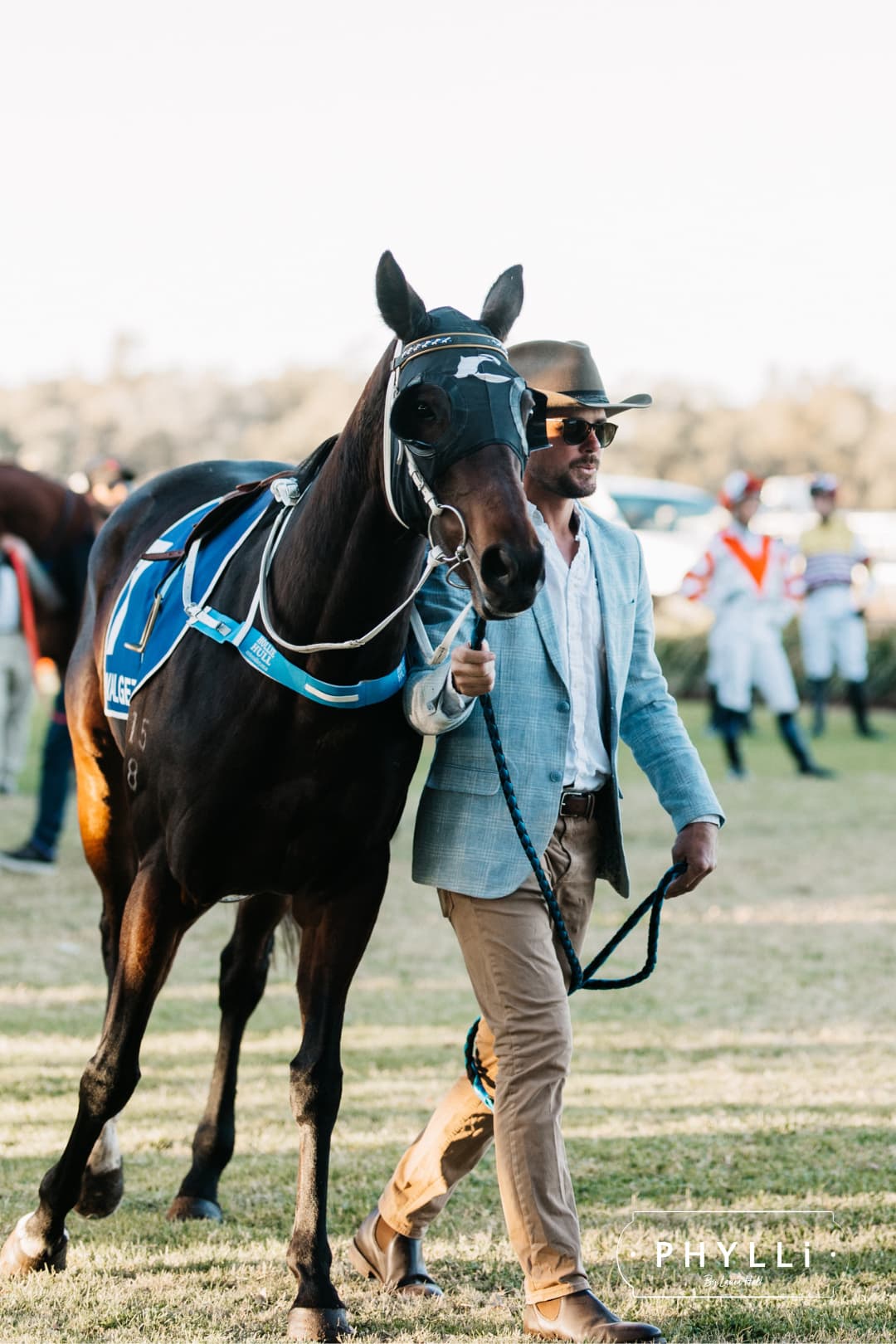 Man wearing a PHYLLi Designs felt hat while leading a racehorse in a racetrack parade ring.