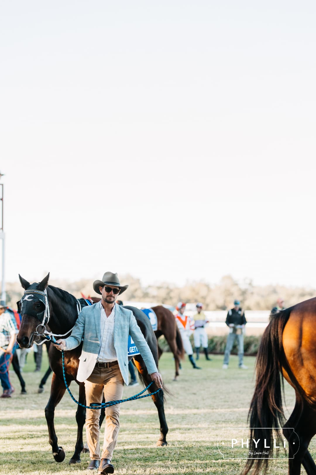 Man wearing a PHYLLi Designs felt cowboy hat while leading a racehorse in a racetrack parade ring.