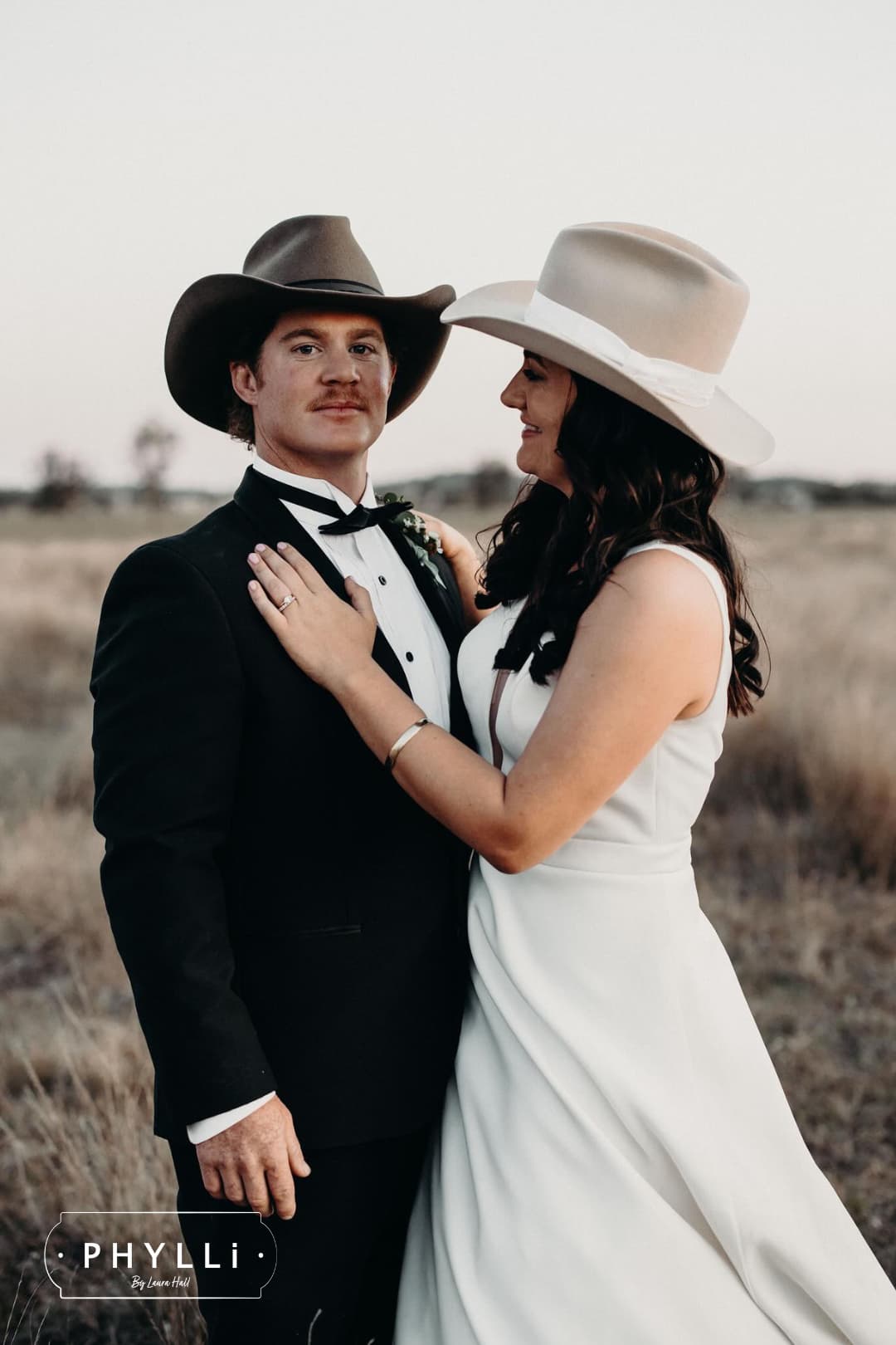 Groom wearing the midnight cowboy felt hat by PHYLLi Designs with bride wearing a beige cowgirl hat in a rural paddock wedding setting.