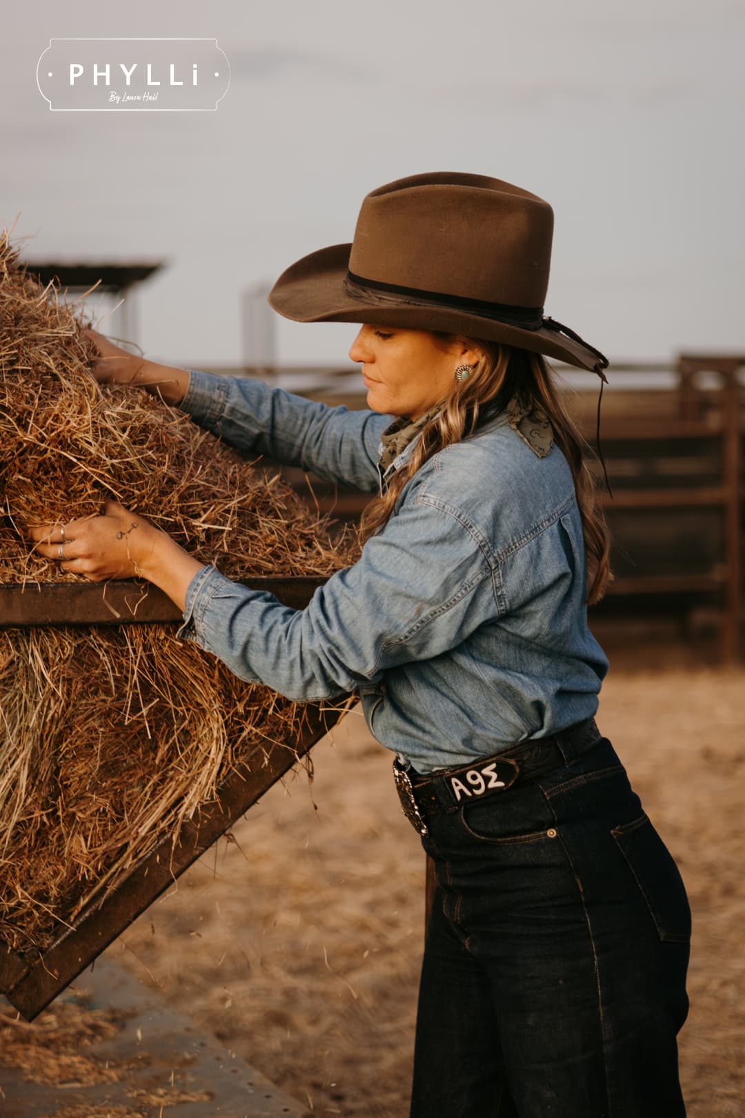 Woman wearing the Midnight Cowboy dirty olive felt cowboy hat by PHYLLi Designs with buffalo leather tieback hat band while feeding hay in cattle yards.