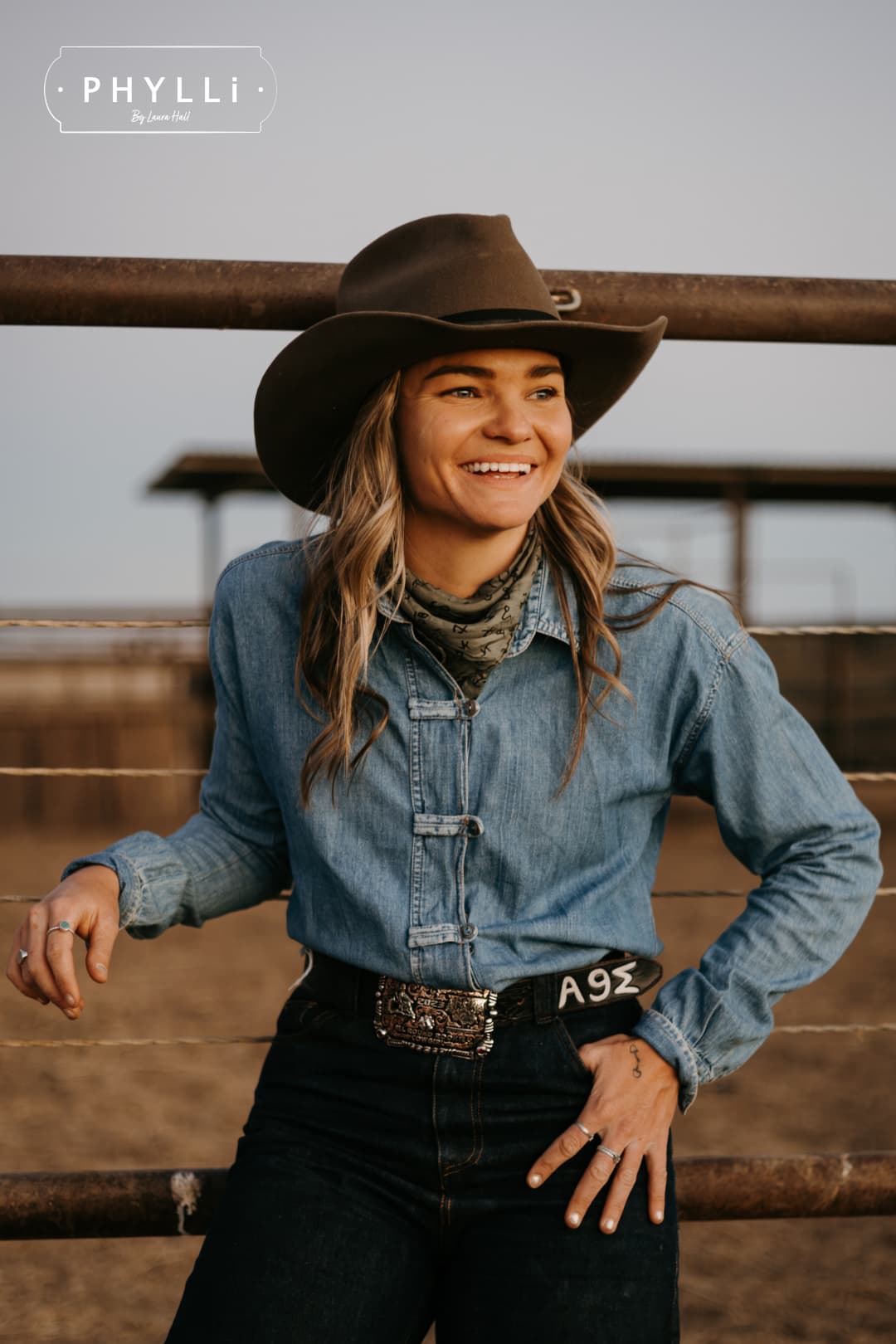 Woman wearing the Midnight Cowboy dirty olive felt cowboy hat by PHYLLi Designs with black Japanese grosgrain ribbon band smiling in rural cattle yards.