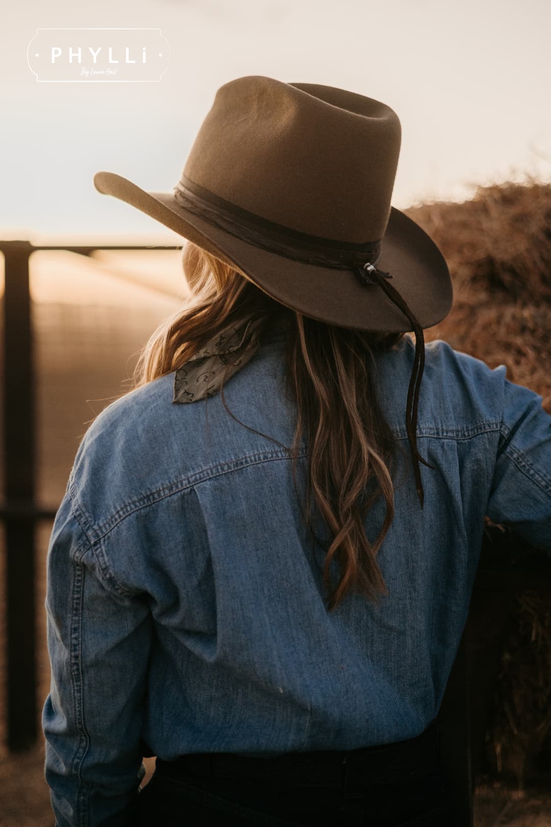 Back profile woman wearing the Midnight Cowboy dirty olive felt cowboy hat by PHYLLi Designs with buffalo leather tieback hat band standing in next to hay bales at sunset.