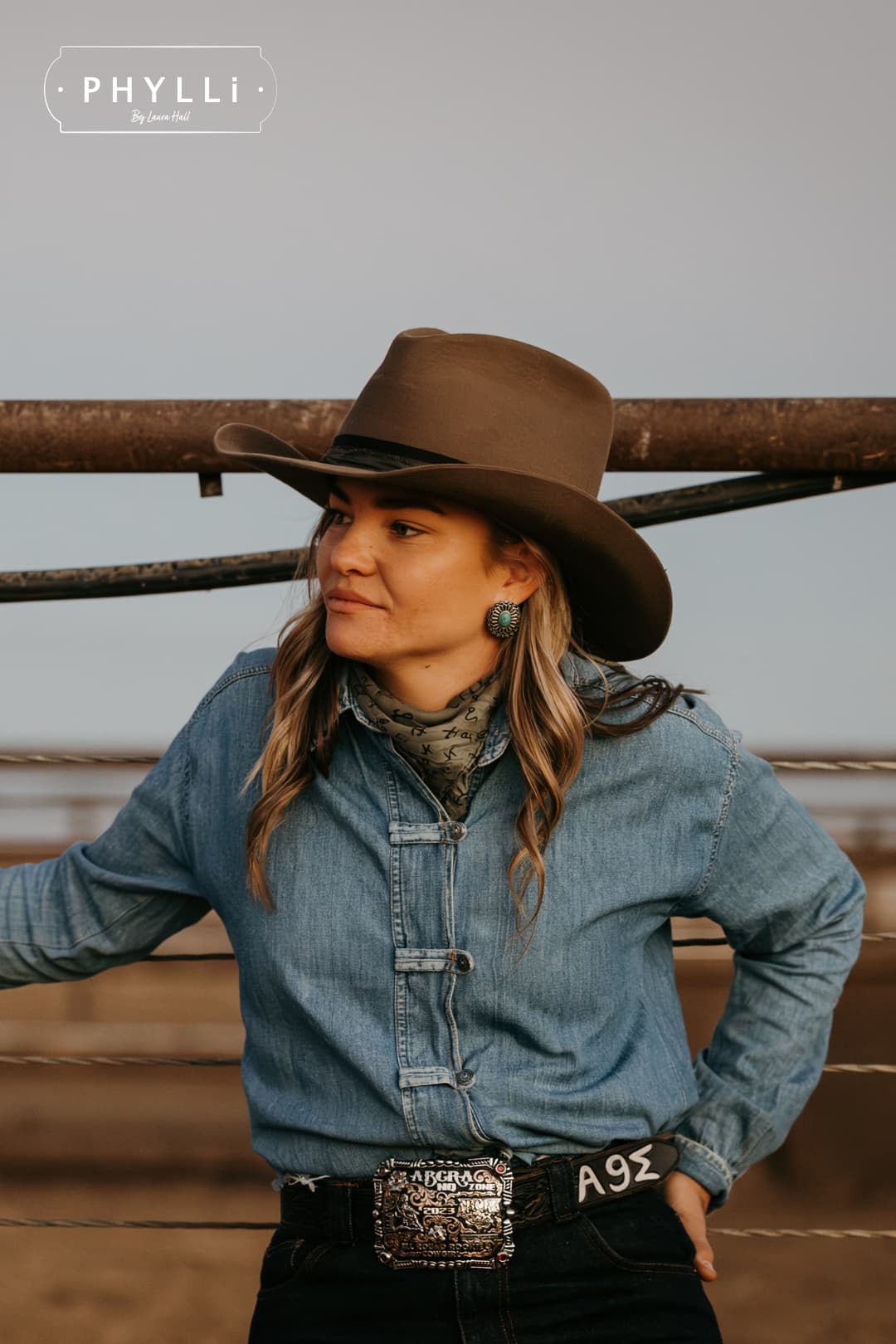 Woman wearing the Midnight Cowboy dirty olive felt cowboy hat by PHYLLi Designs with black Japanese grosgrain ribbon band and midnight sari silk trim leaning on cattle yard rails at sunset.
