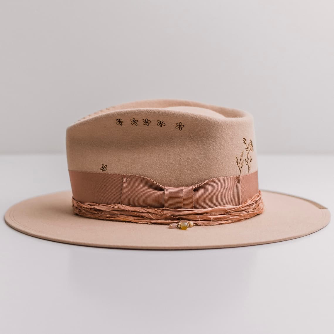 Left side view of the sand felt hat Miley by PHYLLi Designs with dirty rose grosgrain band and burnt floral crown branding.