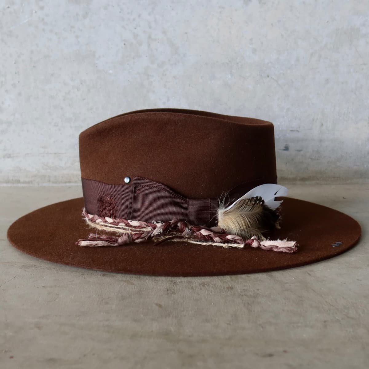 Left side view of the Chocolate felt hat Palmer Limited Edition by PHYLLi Designs with distressed Japanese grosgrain band, plaited sari silk trim and fallen feather bunch.