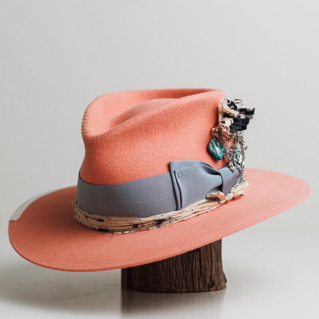 Front left angle of the Pāua peach felt hat by PHYLLi Designs with cornflower grosgrain bow band and silver chunky hat pin with charms and sari silk tassels.
