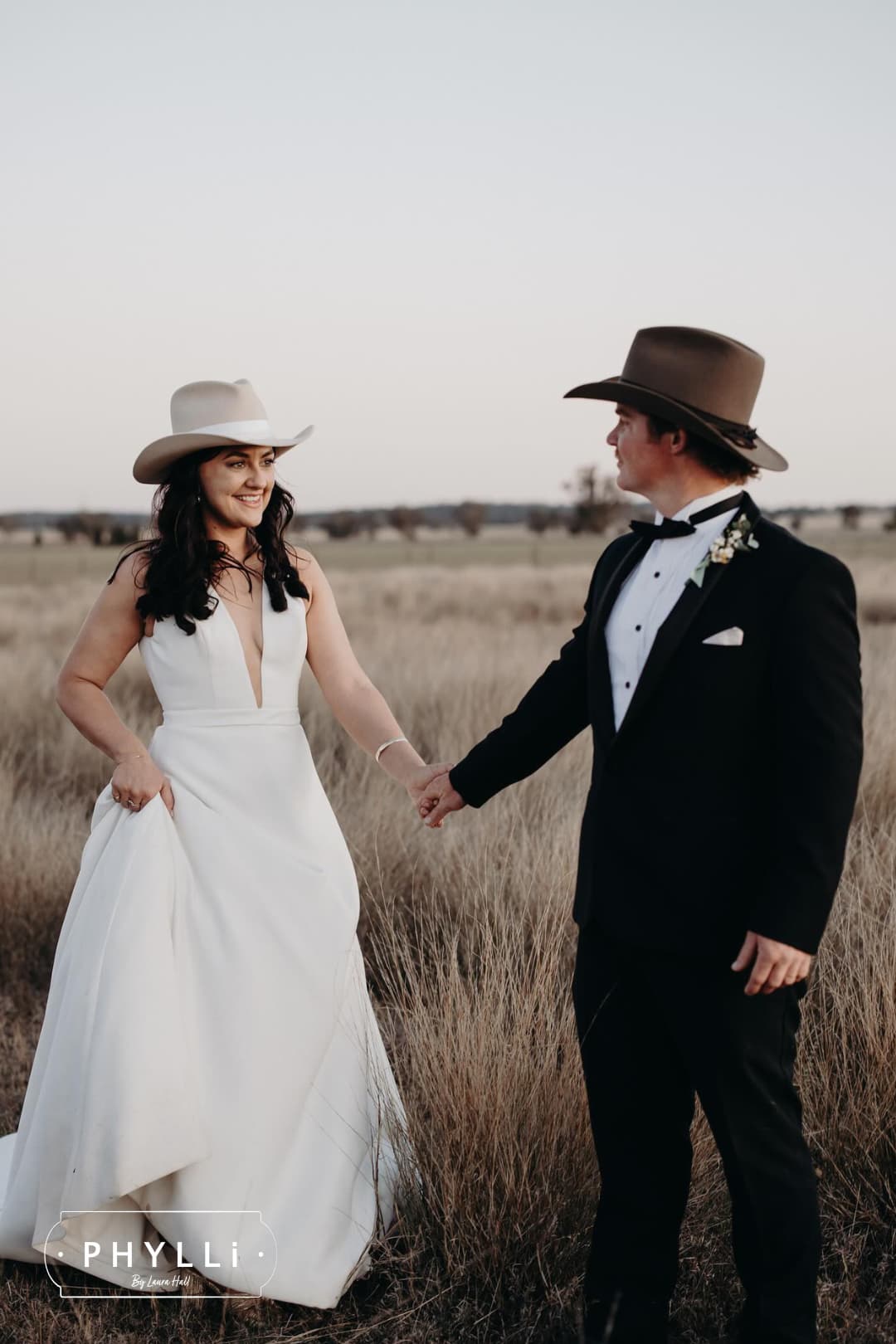 Bride and groom holding hands in an open field wearing beige and dirty olive felt wedding hats by PHYLLi Designs.