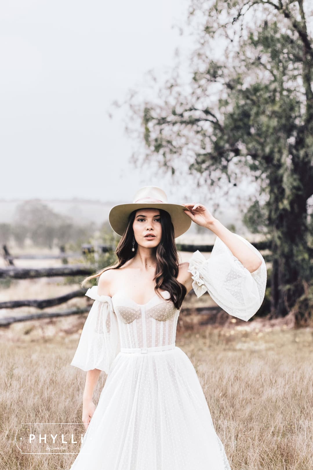 Bride wearing a beige felt bridal hat by PHYLLi Designs with a flowing white gown in a countryside wedding setting.