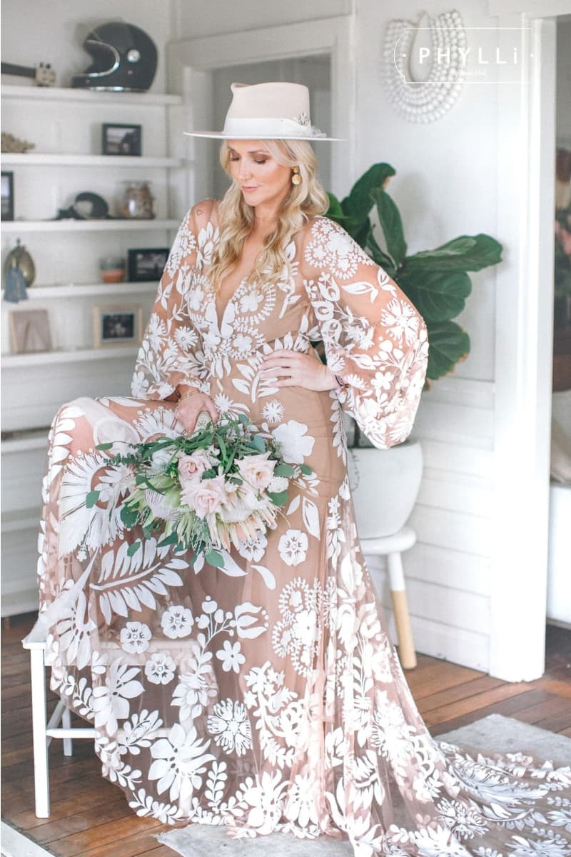 Bride standing indoors wearing a cream bridal felt hat by PHYLLi Designs with floral embroidered wedding gown and bouquet.