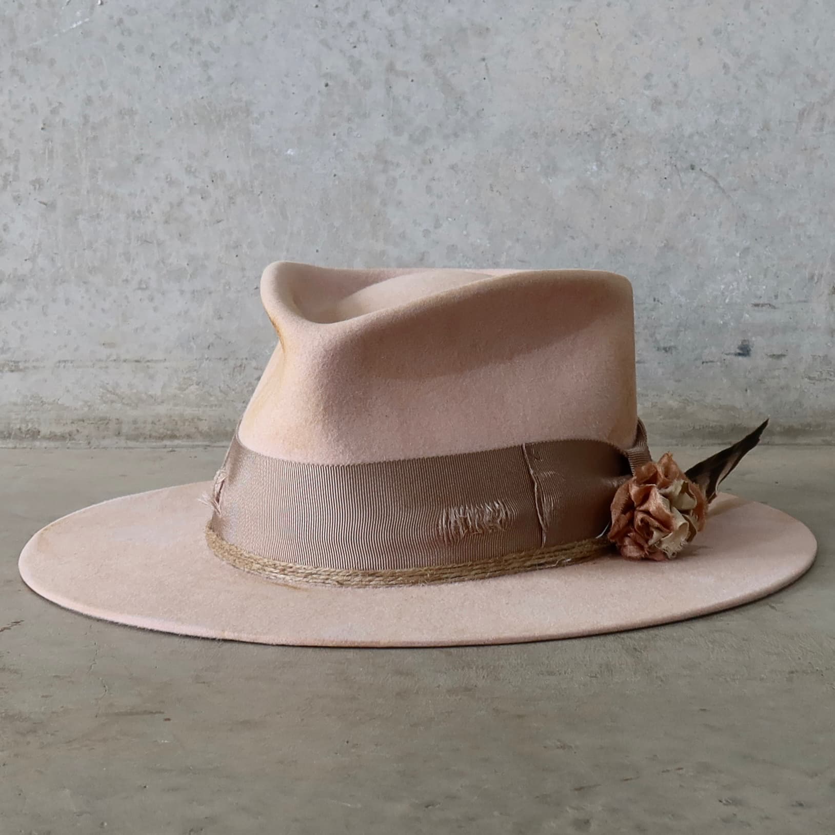Front left angle of the baby pink distressed felt hat Pink Bloom by PHYLLi Designs with taupe grosgrain bow band, twine wraps and sari silk lapel bloom.