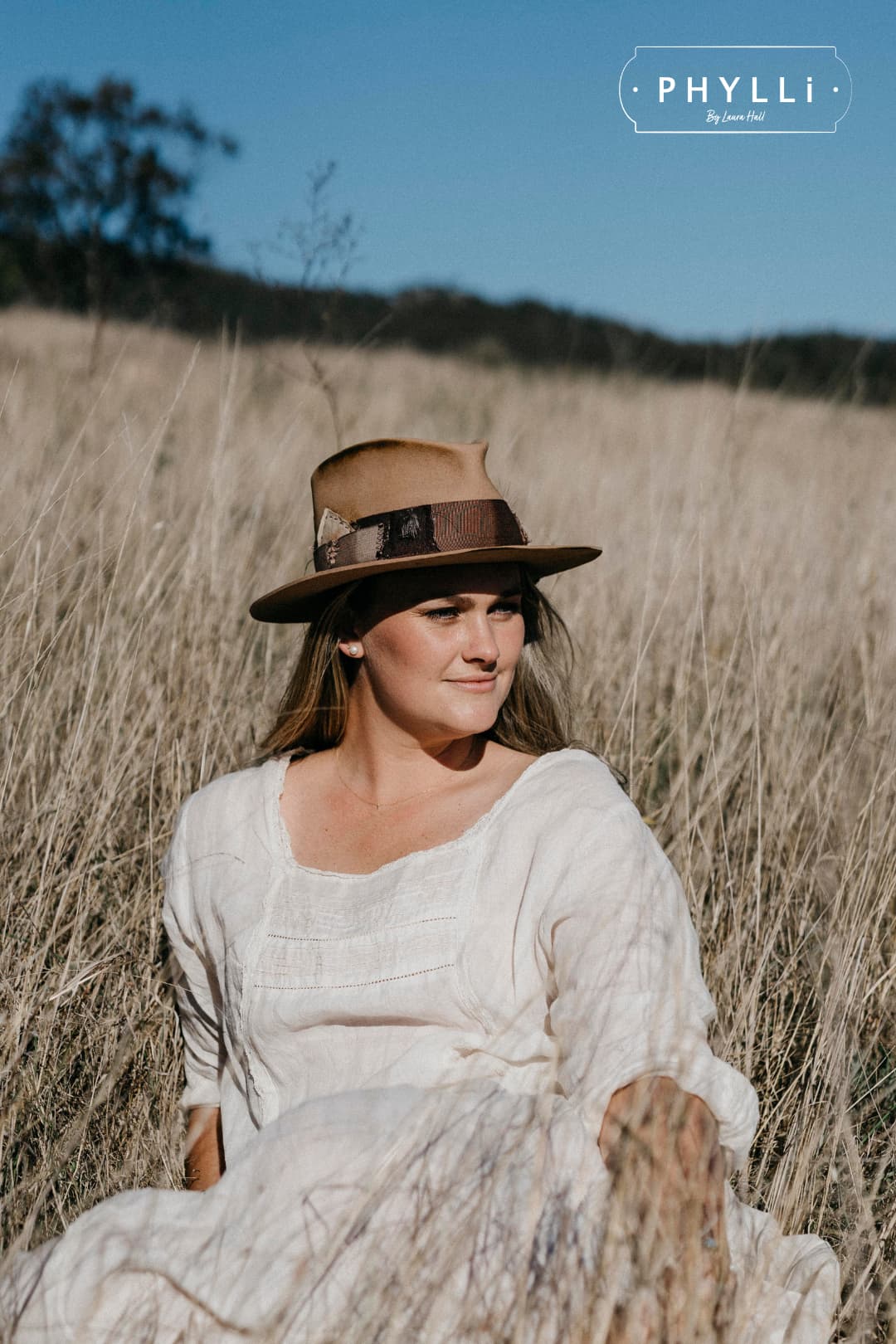 PHYLLi founder Laura Hall wearing the Roadie camel felt hat seated in tall grass.